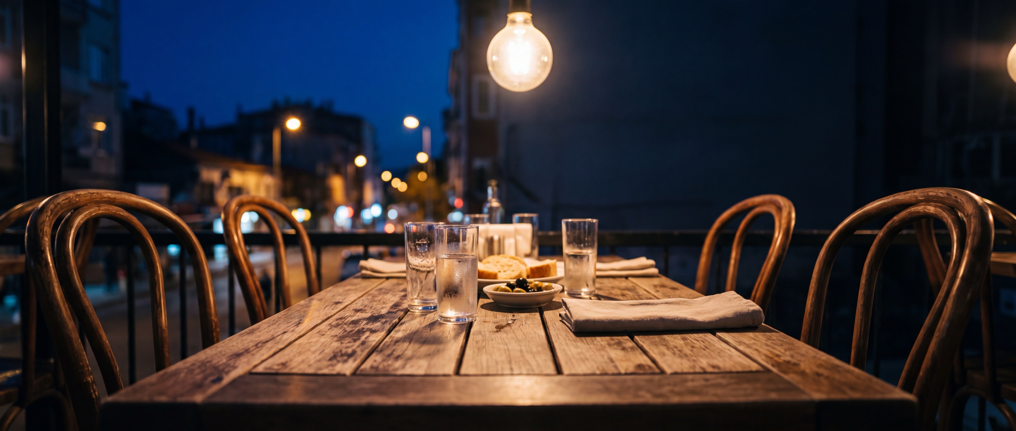 An empty outdoor table set at night — water glasses, bread, olives, a napkin, string lights overhead, a blurred street behind it.